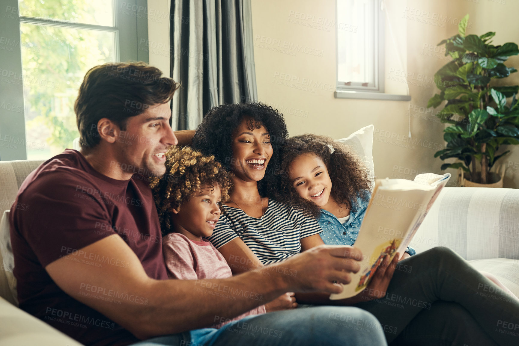 Buy stock photo Shot of a beautiful young family of four reading a story book together while relaxing on a sofa at home