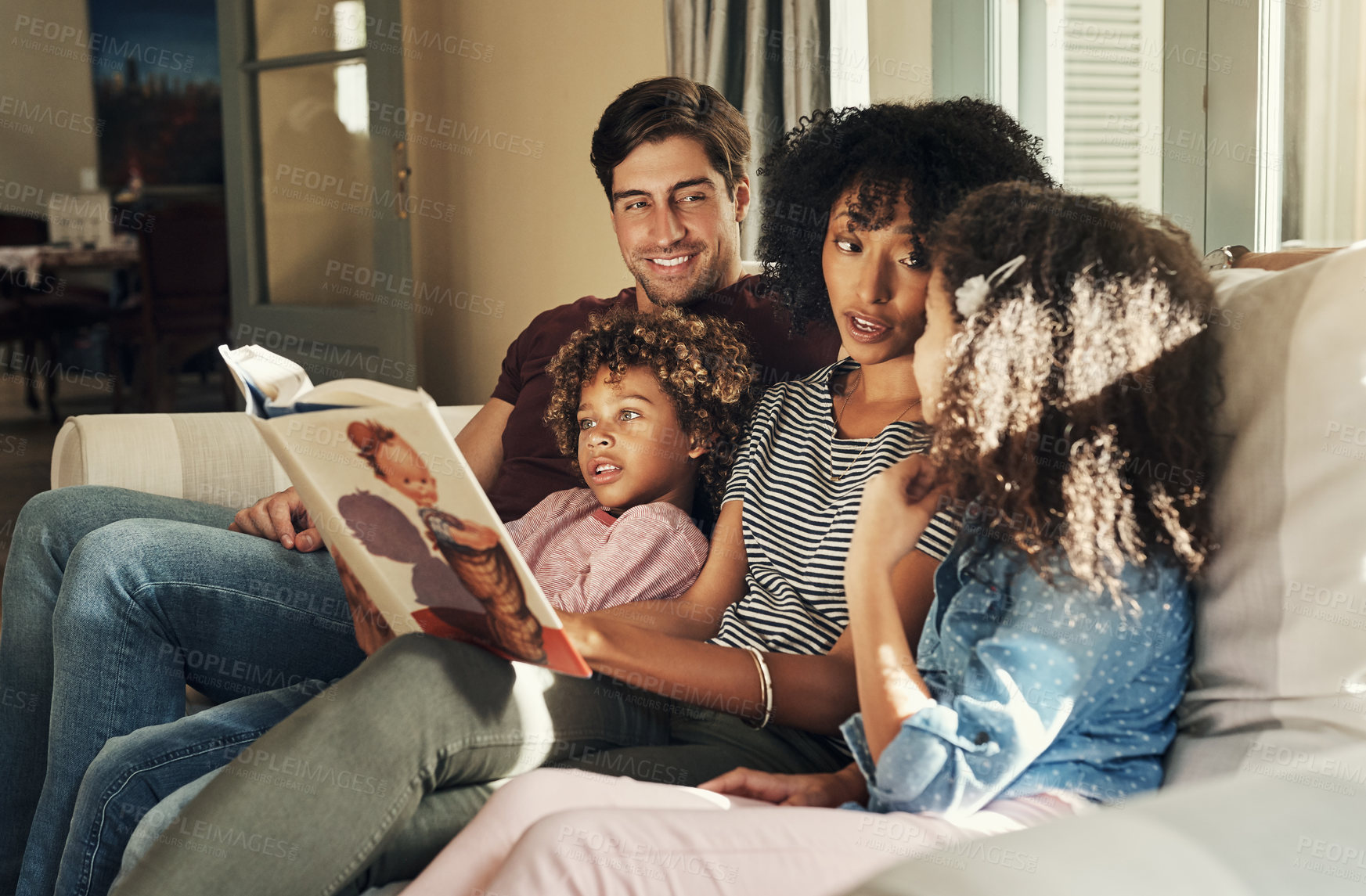 Buy stock photo Shot of a beautiful young family of four reading a story book together while relaxing on a sofa at home