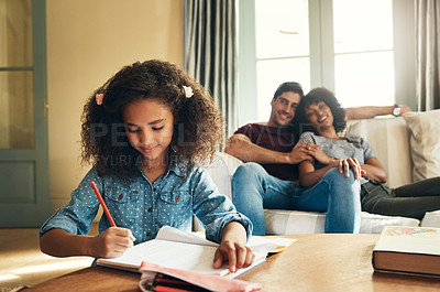 Buy stock photo Shot of an adorable little girl doing her homework with her parents proudly watching in the background at home