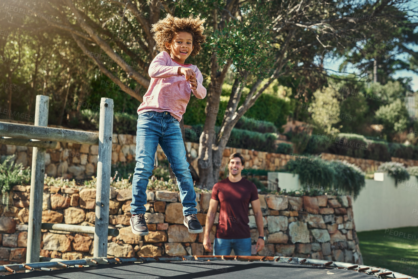 Buy stock photo Shot of an adorable little boy jumping on a trampoline with his father watching closely in the background