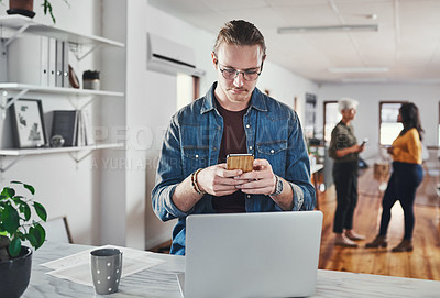 Buy stock photo Cropped shot of a handsome young businessman checking his text messages while working on a laptop in the office