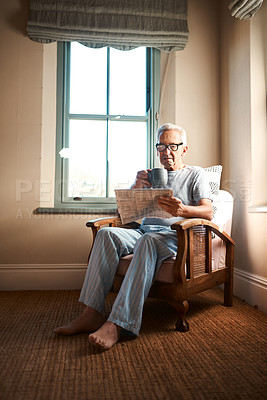 Buy stock photo Full length shot of a senior man sitting and reading a newspaper while holding a cup of coffee at home