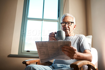 Buy stock photo Cropped shot of a senior man sitting and reading a newspaper while holding a cup of coffee at home