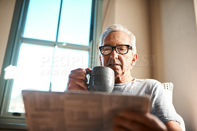 Buy stock photo Cropped shot of a senior man sitting and reading a newspaper while holding a cup of coffee at home