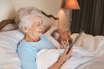 Buy stock photo Cropped shot of a senior woman sitting in bed and using a tablet while her husband sleeps beside her