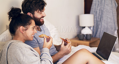 Buy stock photo Shot of a happy young couple using a laptop and eating pizza while relaxing on the bed at home