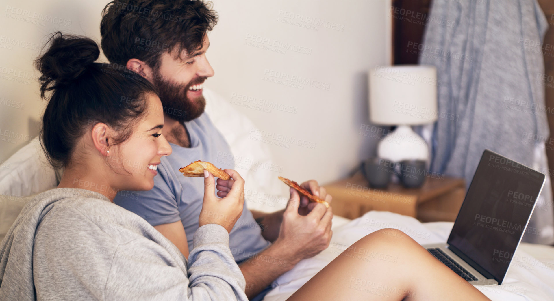 Buy stock photo Shot of a happy young couple using a laptop and eating pizza while relaxing on the bed at home