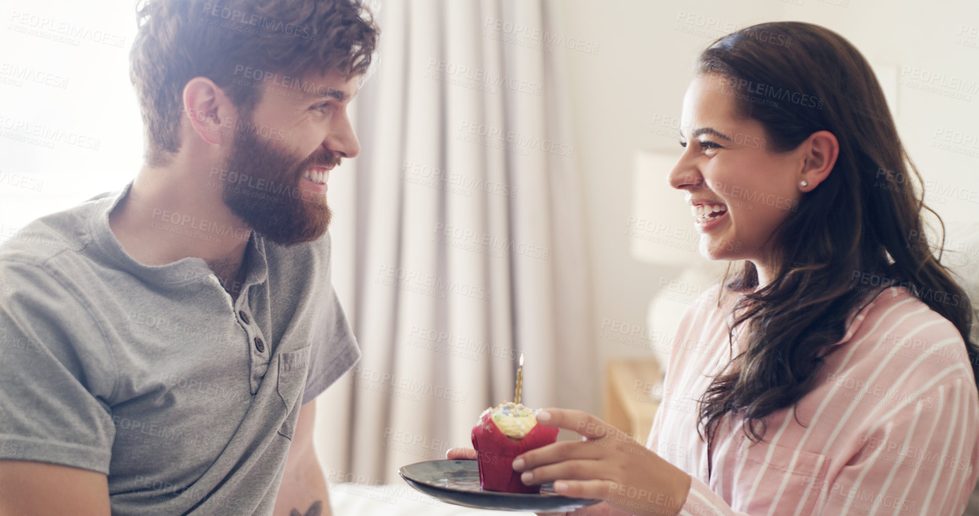 Buy stock photo Shot of a young man surprising his wife with a cupcake in bed at home