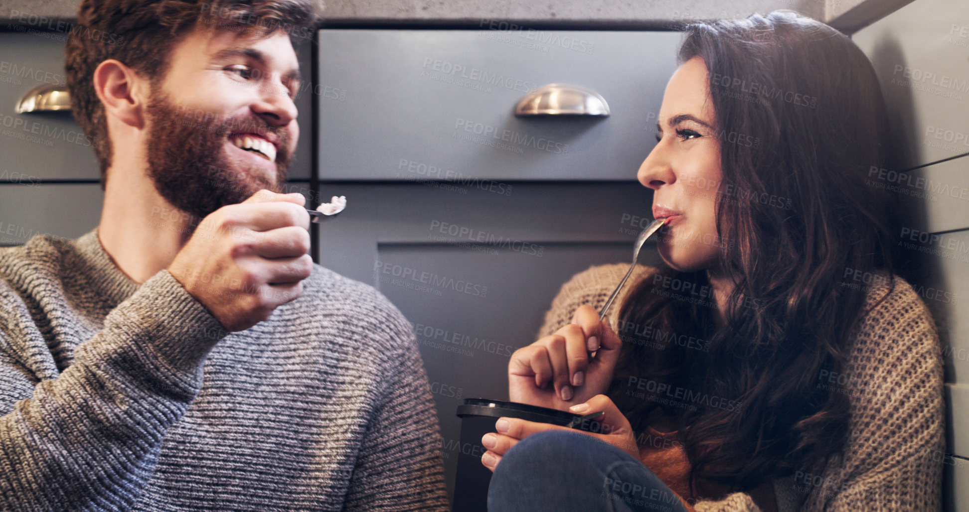 Buy stock photo Shot of a happy young couple sharing a tub of ice cream in their kitchen at home