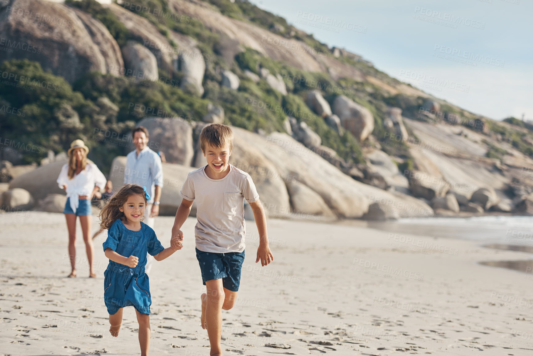 Buy stock photo Cropped shot of two happy siblings holding hands and running along the beach together while their parents watch