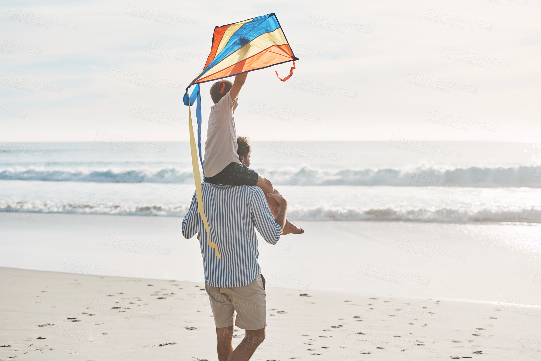 Buy stock photo Rearview shot of a young boy being carried on his father's shoulders and holding a kite on the beach