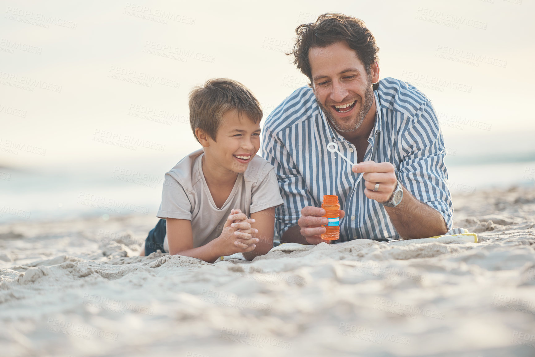 Buy stock photo Full length shot of a playful mature father lying on the beach and blowing bubbles with his son
