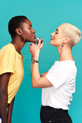 Buy stock photo Studio shot of a young woman putting lipstick on her friend against a turquoise background