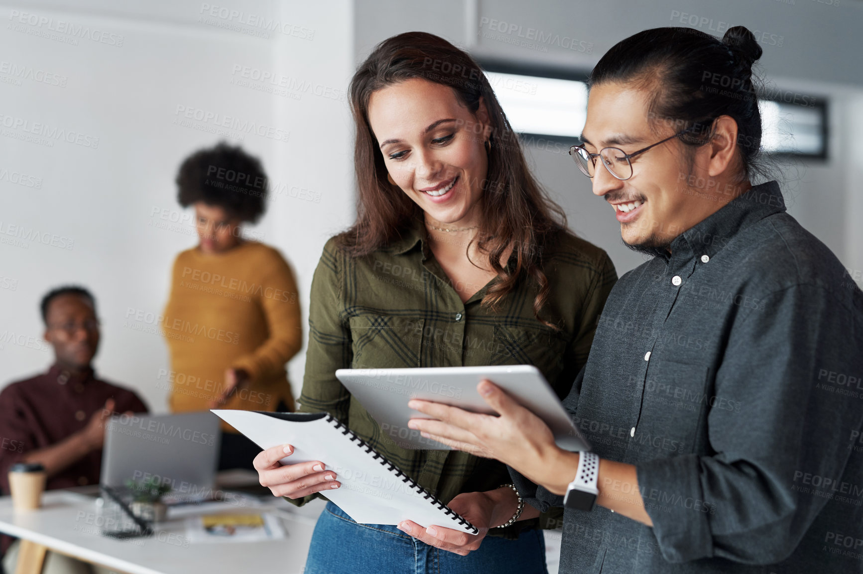 Buy stock photo Shot of two young businesspeople using a digital tablet together at work with their colleagues in the background