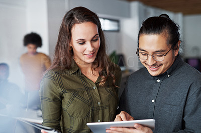 Buy stock photo Shot of two young businesspeople using a digital tablet together at work with their colleagues in the background