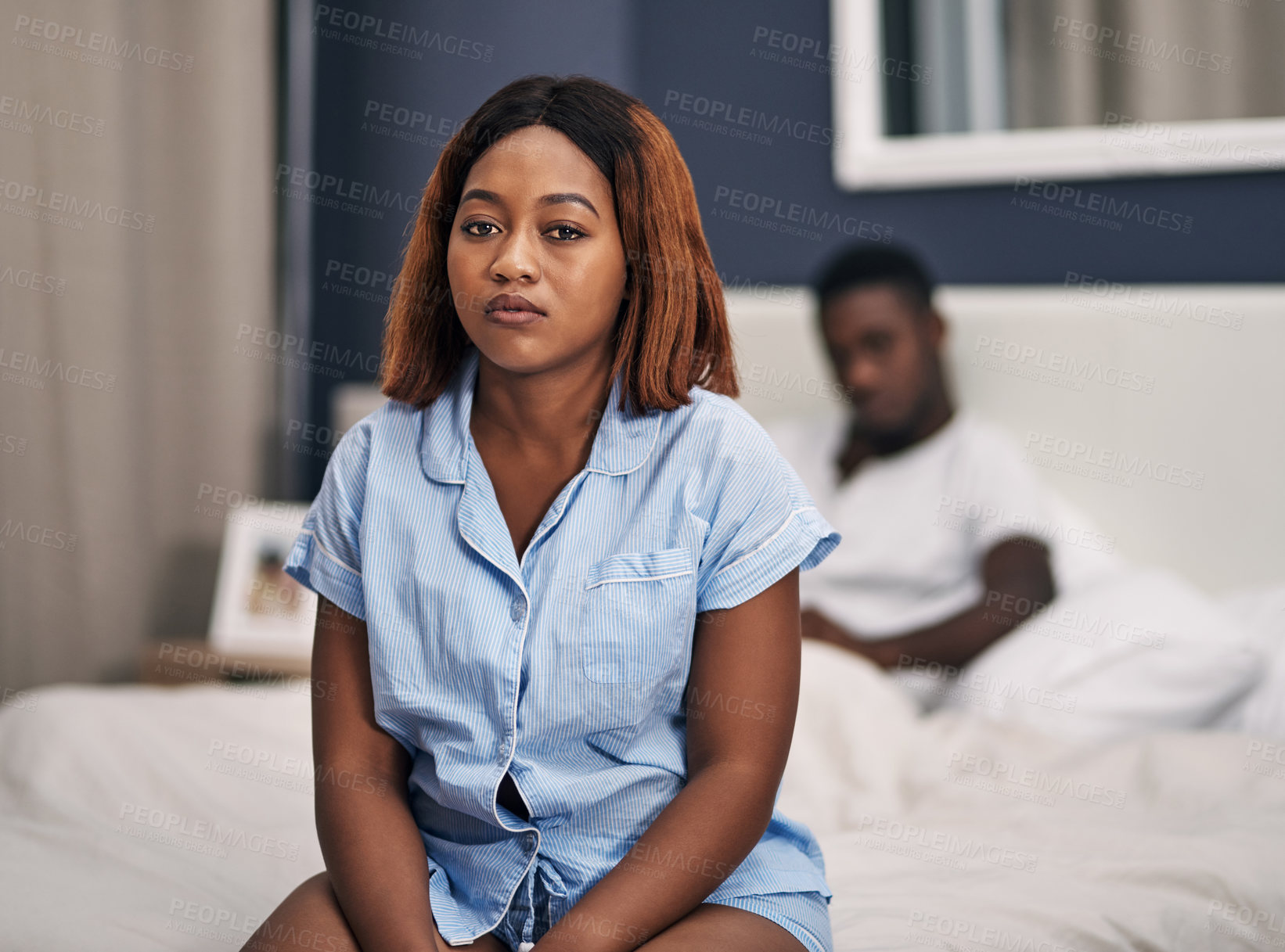 Buy stock photo Cropped portrait of an attractive young woman looking depressed while sitting on the edge of her bed with her husband in the background