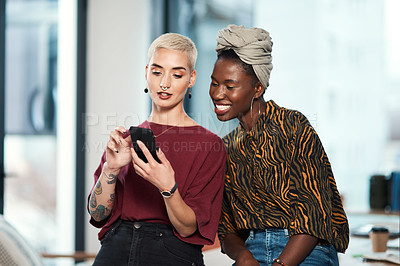 Buy stock photo Cropped shot of two attractive young business colleagues sitting in the office together and using a cellphone