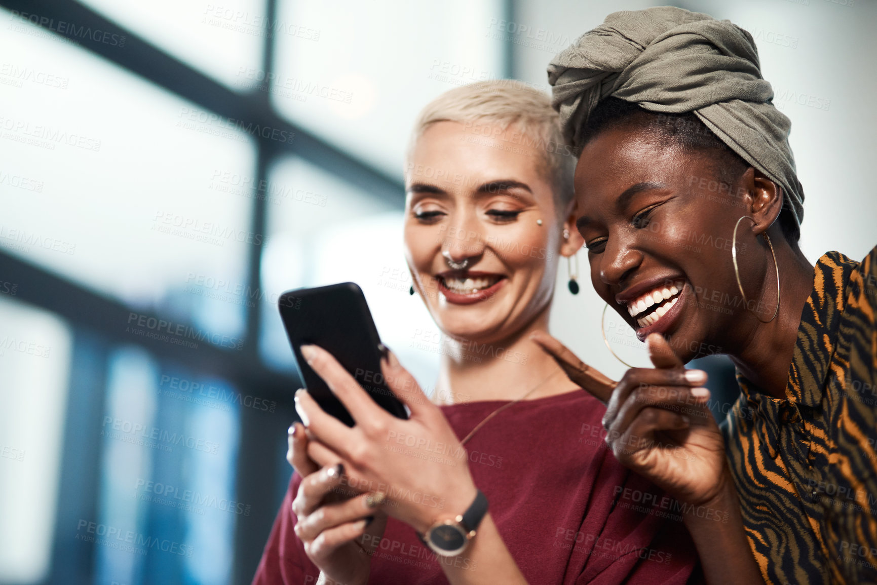 Buy stock photo Cropped shot of two attractive young business colleagues standing in the office together and laughing while using a cellphone