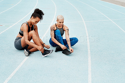 Buy stock photo Full length shot of two attractive young athletes sitting together and tying their shoelaces before running on a track