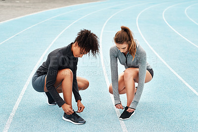 Buy stock photo Full length shot of two attractive young athletes crouching together and tying their shoelaces before running on a track