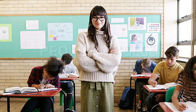 Buy stock photo Portrait of a cheerful young female teacher standing with her arms folded while her students do work inside of a classroom at school