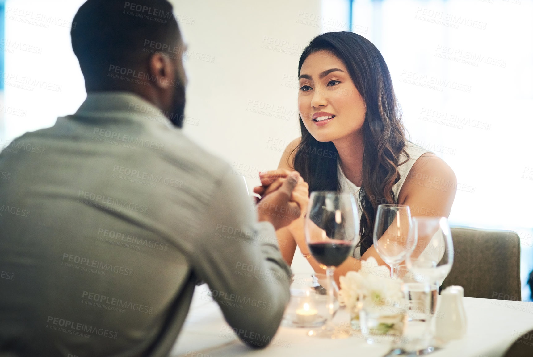 Buy stock photo Couple, holding hands and wine in restaurant with talking, love and romance for valentines day date in night. Black man, asian woman and conversation for bonding, luxury fine dining and celebration