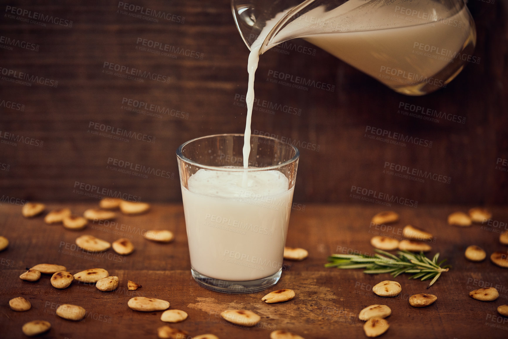 Buy stock photo Shot of a jug pouring of milk into a glass surrounded by nuts on a table