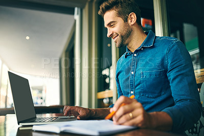 Buy stock photo Cropped shot of a handsome young businessman sitting alone and making notes while using his laptop in a cafe
