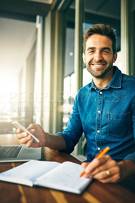 Buy stock photo Cropped portrait of a handsome young businessman sitting alone and making notes while using his cellphone in a cafe
