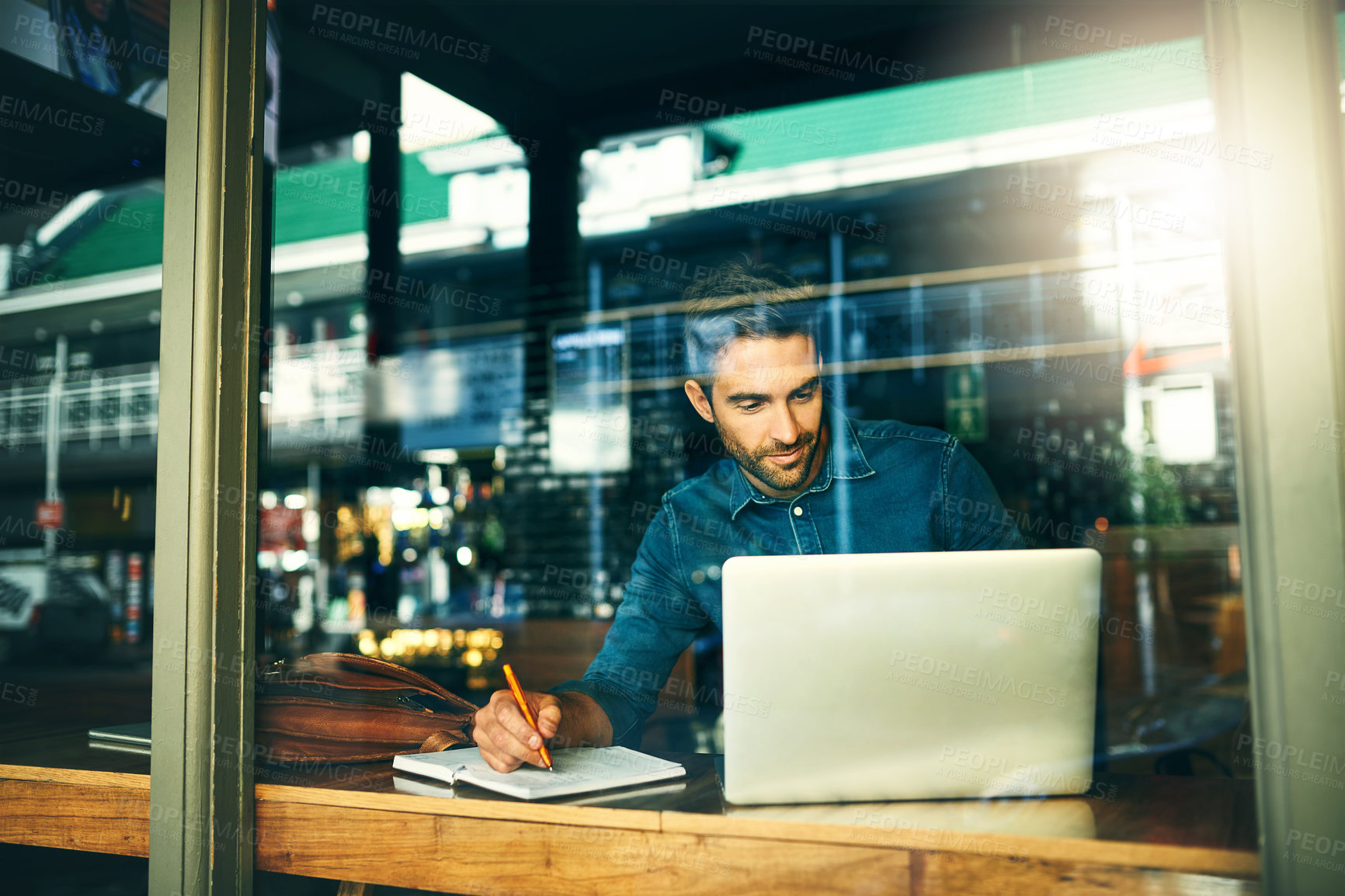 Buy stock photo Cropped shot of a handsome young businessman sitting alone and making notes while using his laptop in a cafe