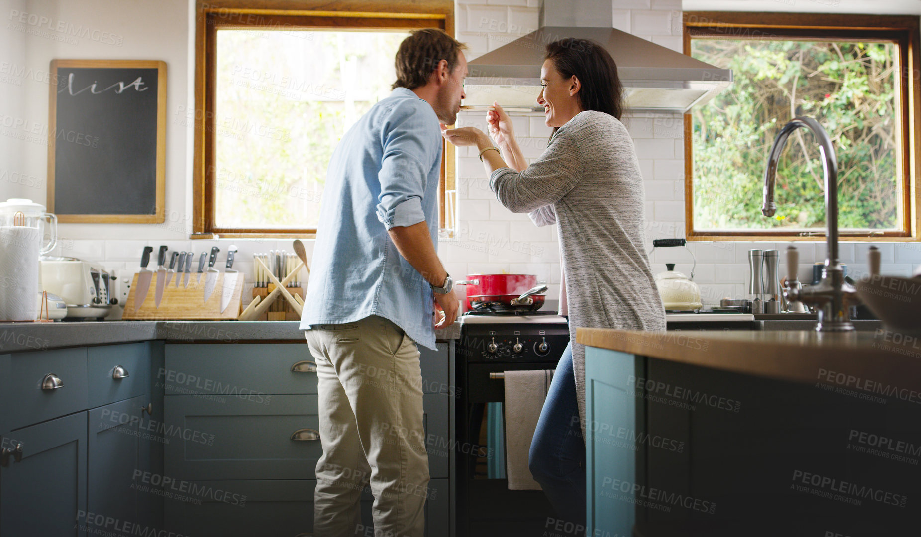 Buy stock photo Cropped shot of an affectionate young woman feeding her husband a spoonful of her food while cooking in their kitchen