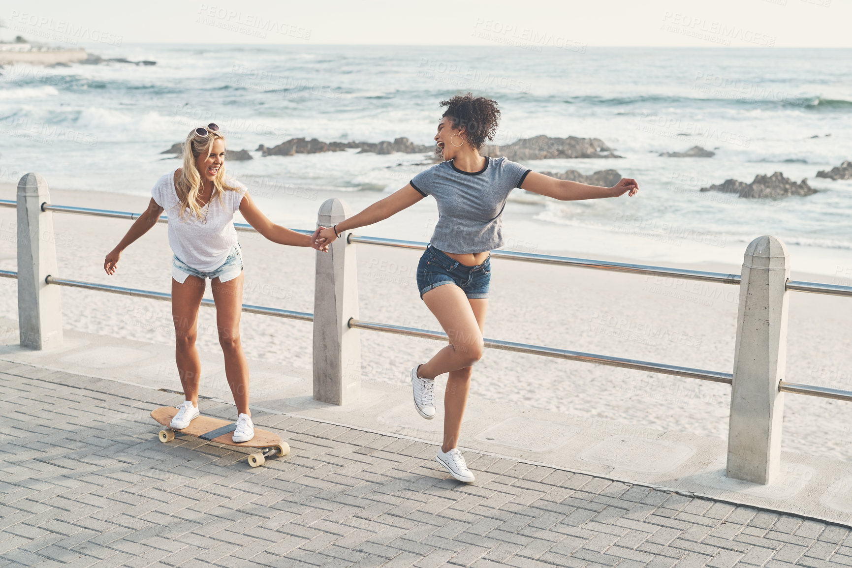 Buy stock photo Shot of a woman pulling her friend on a skateboard while out on the promenade