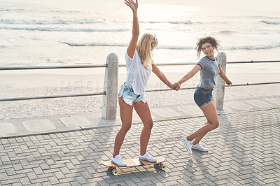 Buy stock photo Shot of a woman pulling her friend on a skateboard while out on the promenade