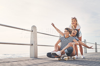 Buy stock photo Shot of a woman pushing her friends on a skateboard while out on the promenade
