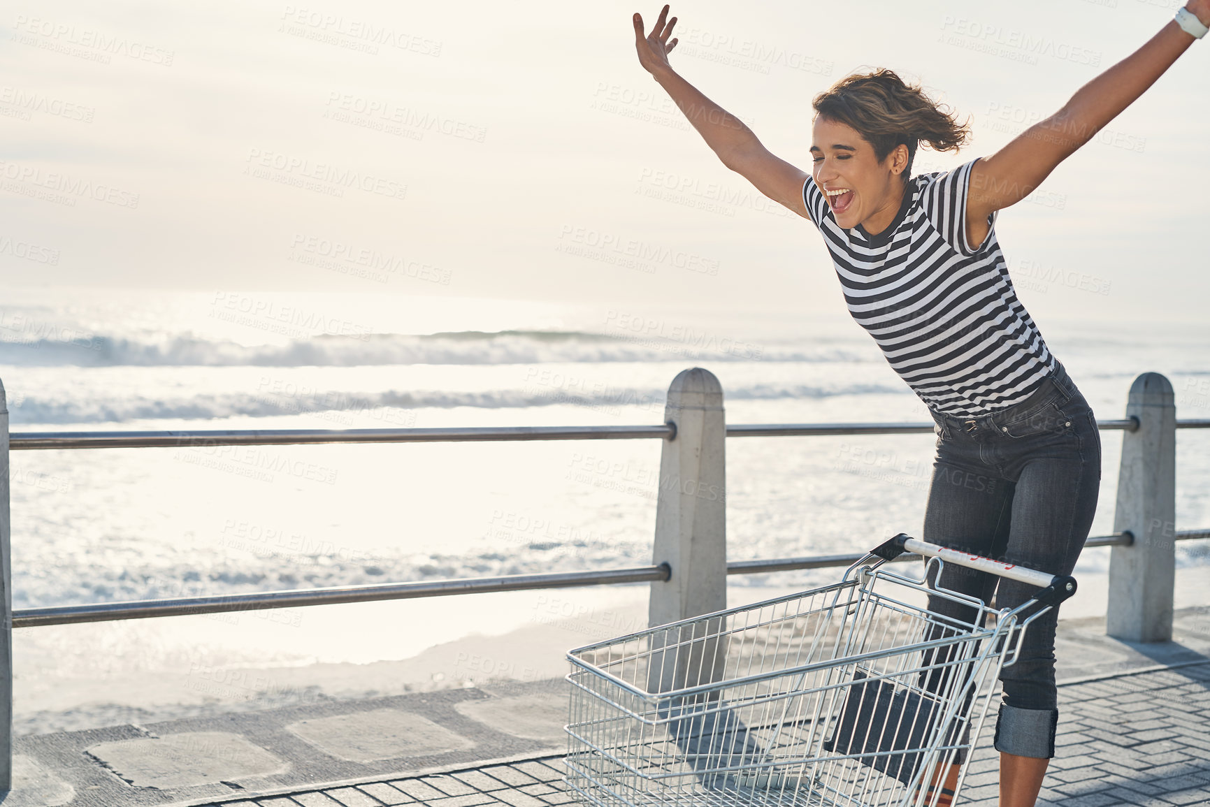 Buy stock photo Shot of a young woman riding a shopping cart on the promenade