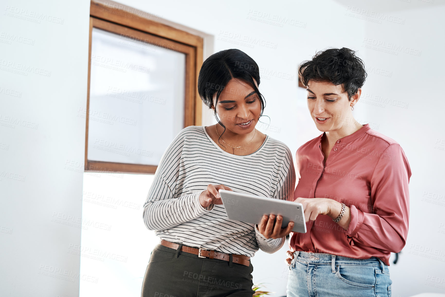 Buy stock photo Cropped shot of two attractive young businesswomen standing together in the office and using a tablet