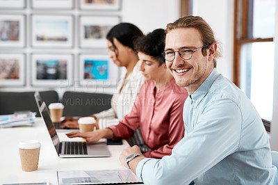Buy stock photo Cropped portrait of a handsome young businessman sitting while his colleagues work behind him in the office