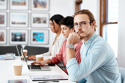 Buy stock photo Cropped portrait of a handsome young businessman sitting and looking contemplative while his colleagues work behind him in the office
