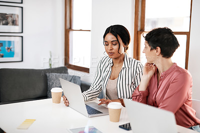 Buy stock photo Cropped shot of two young businesswomen sitting together in the office and using laptops during a meeting
