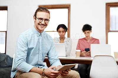 Buy stock photo Cropped portrait of a handsome young businessman sitting and using a tablet while his colleagues work behind him