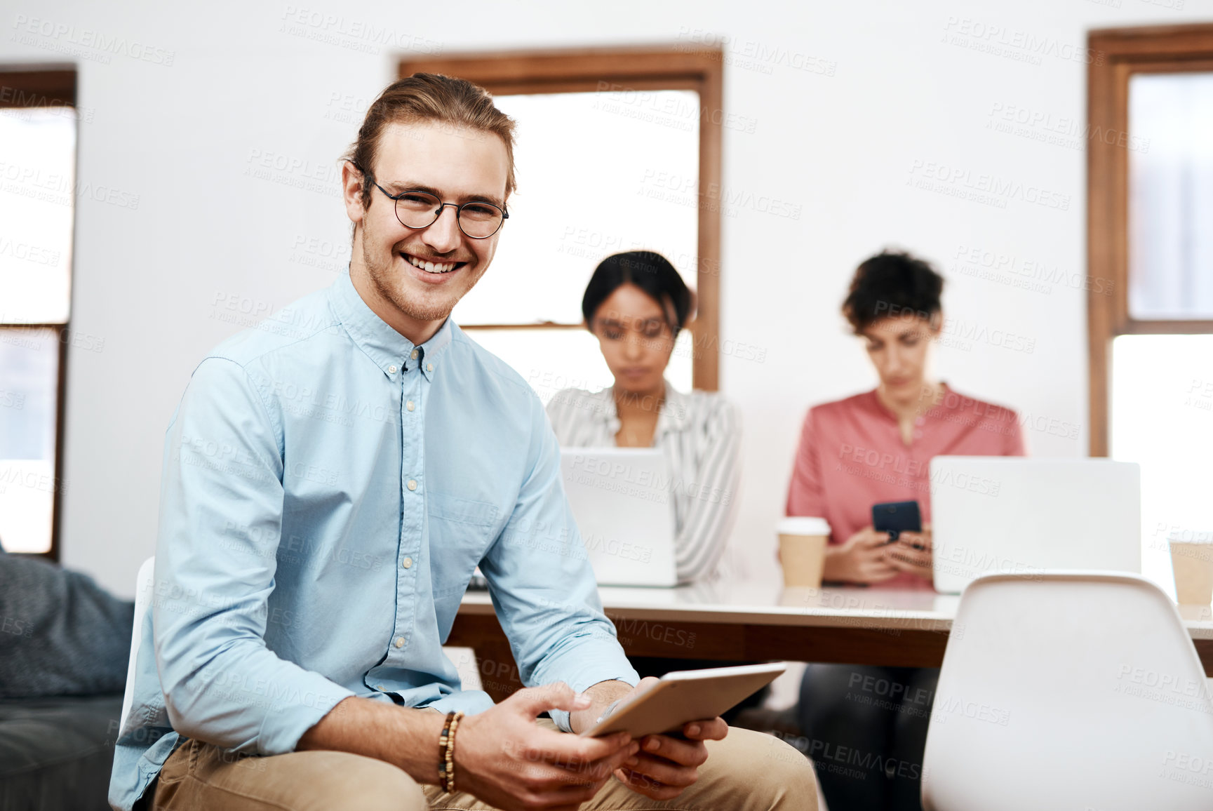 Buy stock photo Cropped portrait of a handsome young businessman sitting and using a tablet while his colleagues work behind him