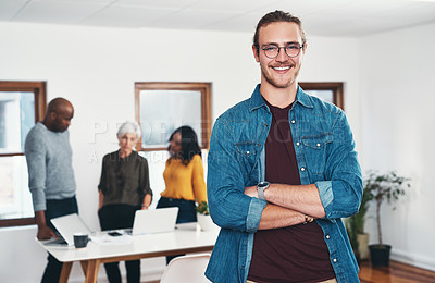 Buy stock photo Portrait of a cheerful young businessman standing with his arms folded and his work colleagues in the background inside of the office