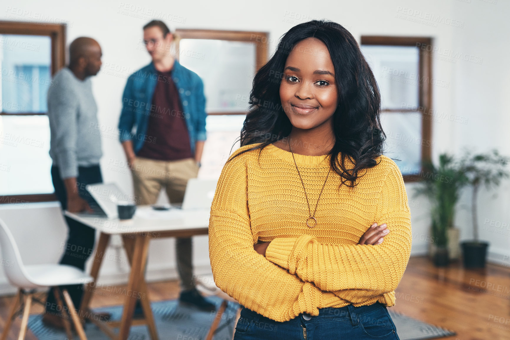 Buy stock photo Portrait of a cheerful young businesswoman standing with her arms folded and her work colleagues in the background inside of the office