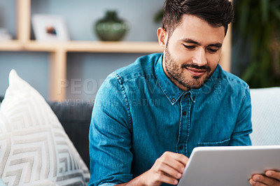 Buy stock photo Cropped shot of a handsome young man looking cheerful while using a digital tablet in his living room at home