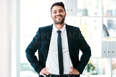 Buy stock photo Cropped portrait of a handsome young businessman smiling while standing with his arms on his waist in a modern office