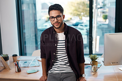 Buy stock photo Cropped portrait of a handsome young businessman wearing spectacles and leaning on his desk while working alone in his office