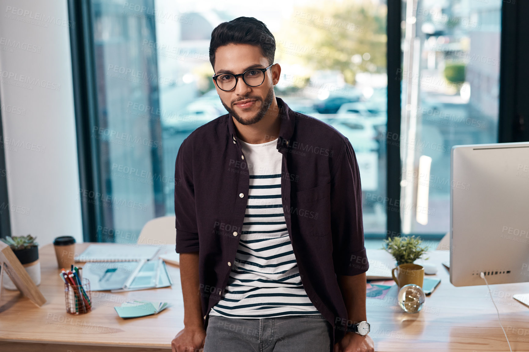 Buy stock photo Cropped portrait of a handsome young businessman wearing spectacles and leaning on his desk while working alone in his office