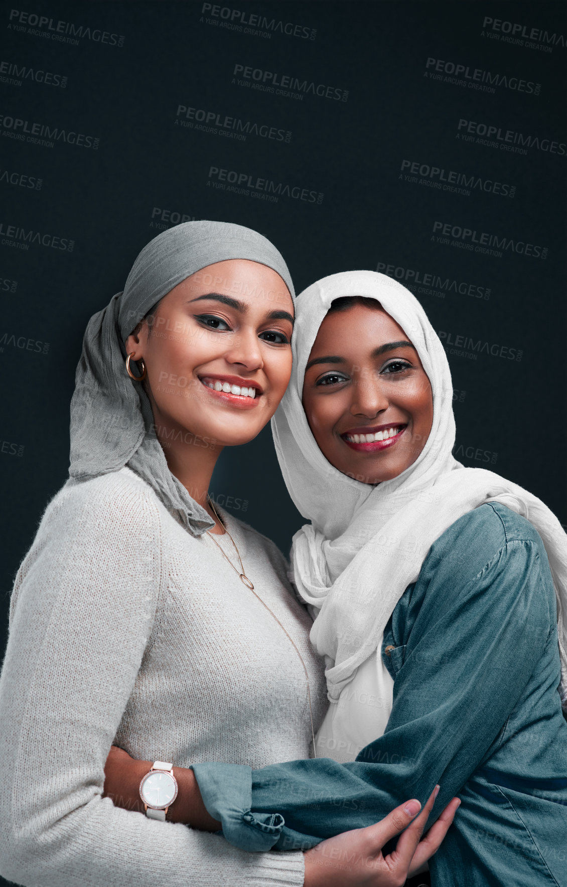 Buy stock photo Cropped shot of two attractive young women wearing hijabs and embracing each other against a black background in the studio