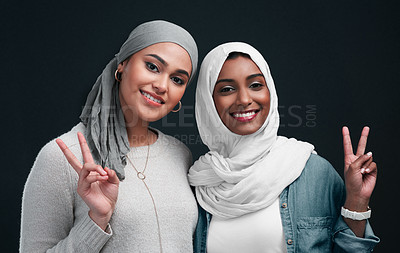 Buy stock photo Cropped shot of two attractive young women wearing hijabs and standing together while showing a peace sign in the studio