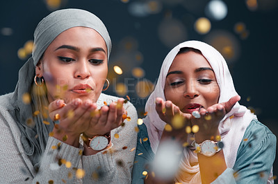 Buy stock photo Cropped shot of two attractive young women wearing hijabs and blowing confetti while standing against a black studio background